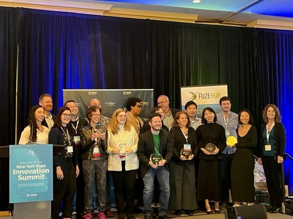 A group of people stand on a stage holding awards and smiling next to a sign for the New York State Innovation Summit.